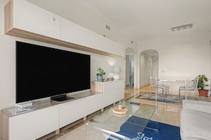 A modern living room with a large flat-screen TV, a glass coffee table, and a dining area with a white table and metal chairs.