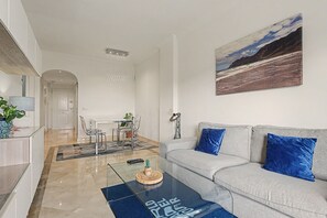A modern living room with a glass coffee table, a grey sofa with blue cushions, a dining area with a table and chairs, and a large landscape painting on the wall.