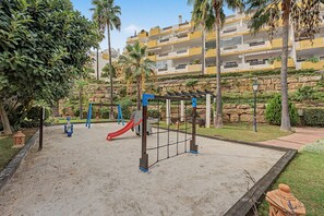 A playground with a slide and swings, surrounded by a landscaped area with palm trees and a stone wall.