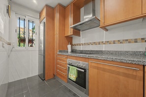 A modern kitchen with wooden cabinets, a stainless steel range hood, a built-in oven, and a window with a view of buildings outside.