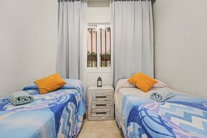 A small, clean room with two beds, each with a patterned bedspread and a single pillow. There is a small wooden nightstand with a lantern between the beds.