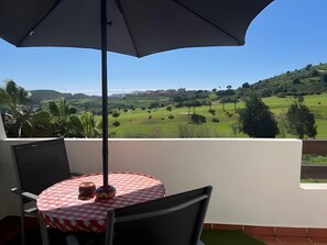 Un balcon avec une table et des chaises, donnant sur un paysage verdoyant, avec des arbres et des bâtiments visibles au loin.