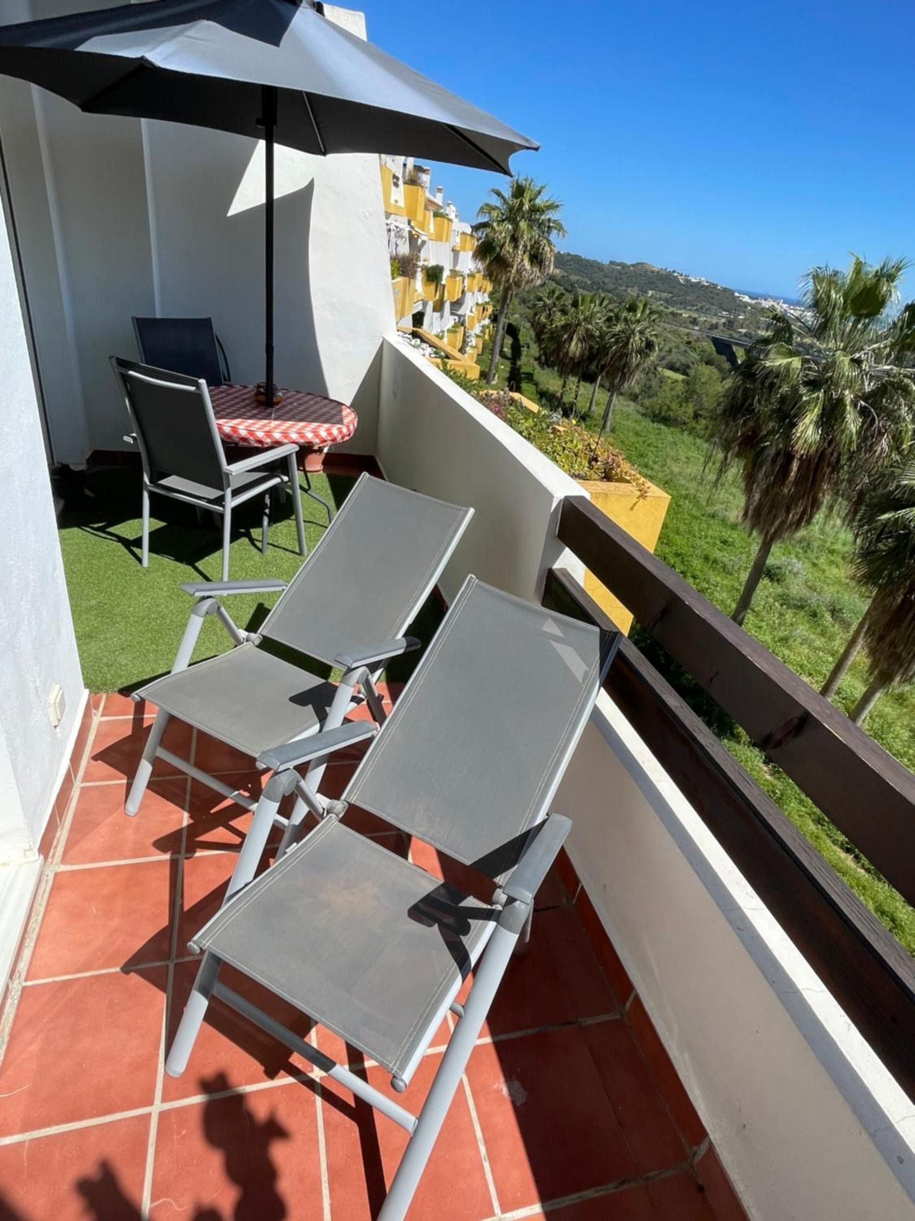 Ein Balkon mit zwei Stühlen und einem Tisch, einem Sonnenschirm und Blick auf einen Hügel mit Palmen.