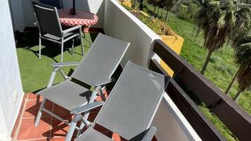 Ein Balkon mit zwei Stühlen und einem Tisch, einem Sonnenschirm und Blick auf einen Hügel mit Palmen.