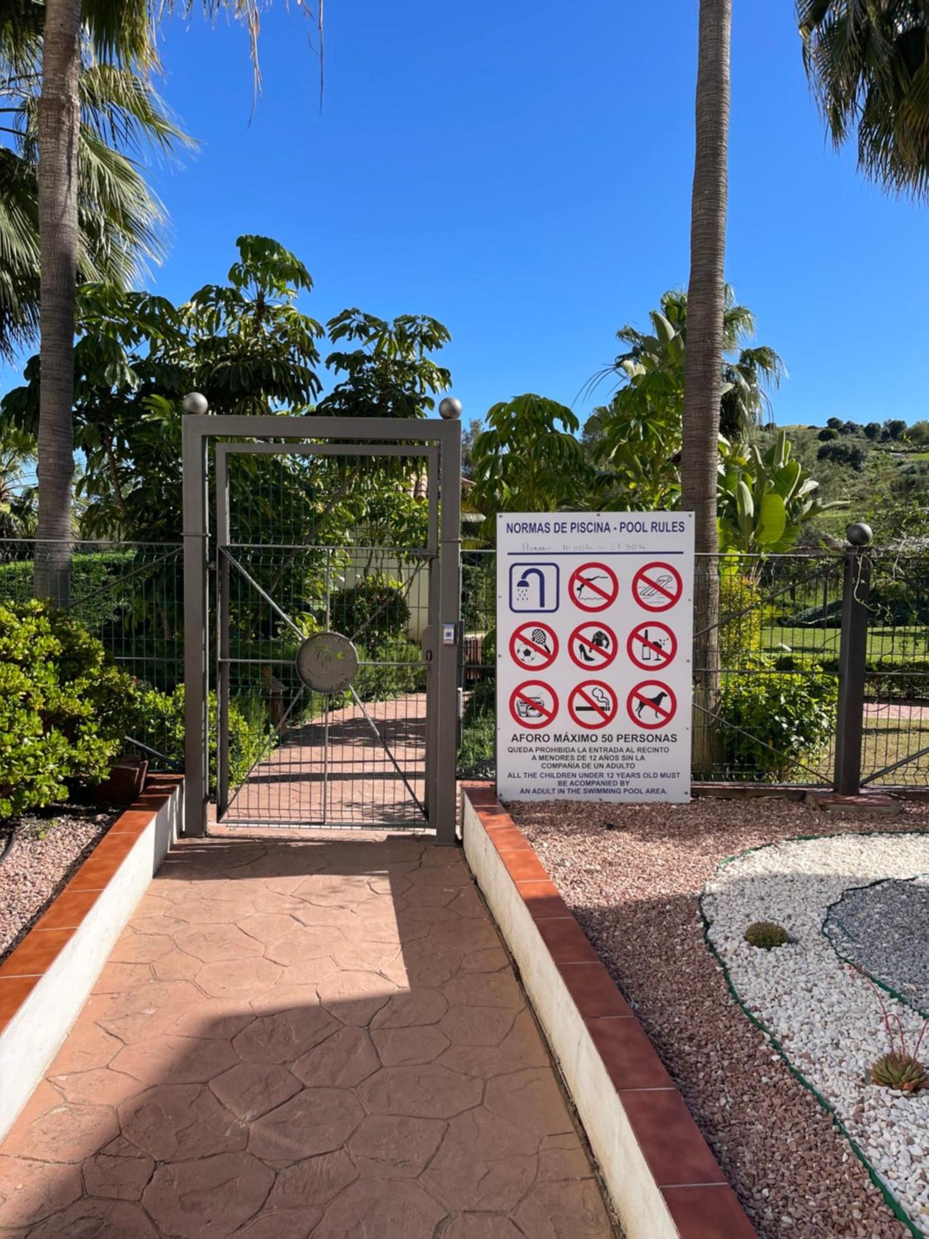 Une entrée fermée par un portail mène à un espace piscine où un panneau affiche les règles d’utilisation de la piscine.