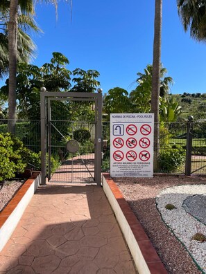Une entrée fermée par un portail mène à un espace piscine où un panneau affiche les règles d’utilisation de la piscine.