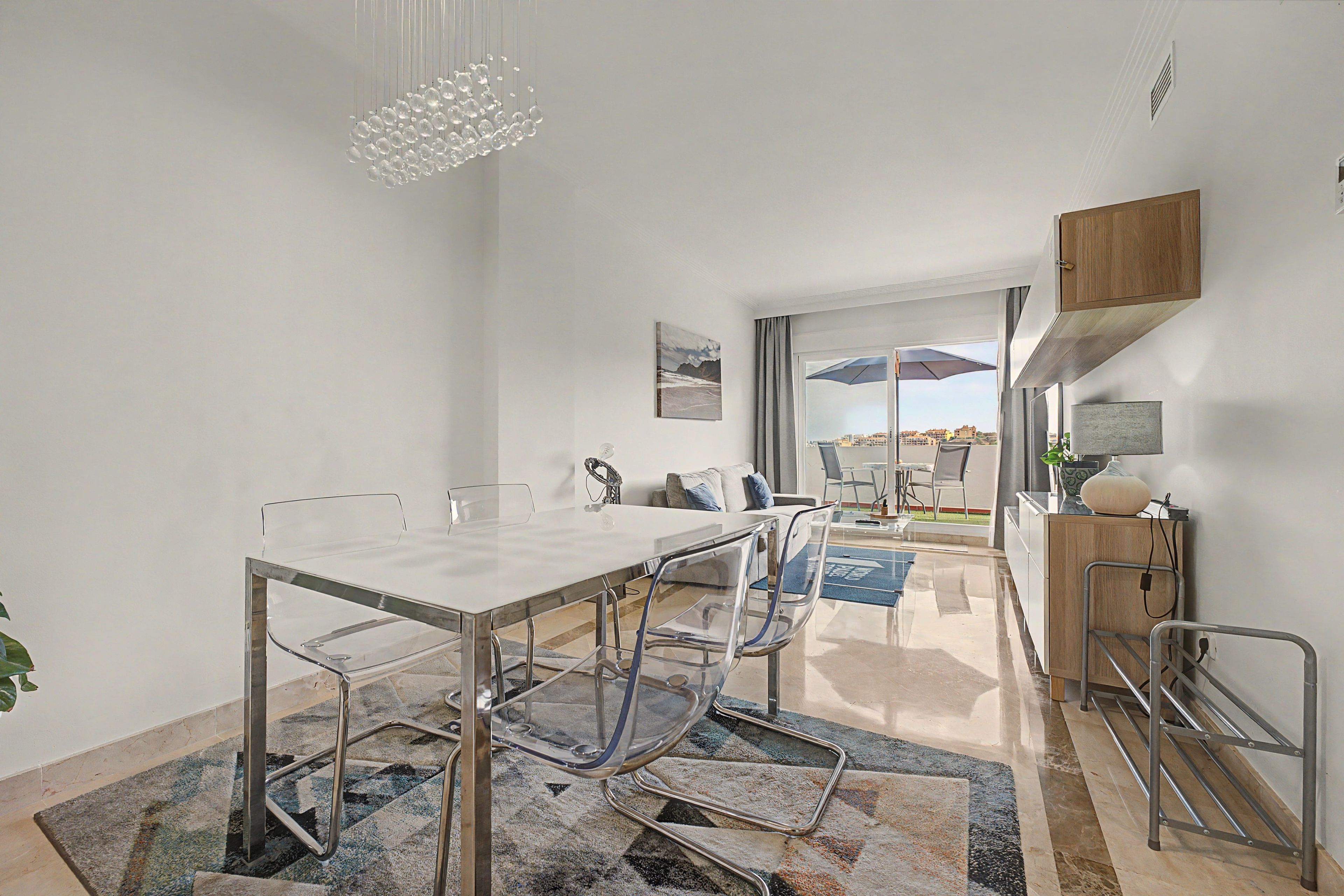 A modern kitchen with a dining area, featuring a glass table, metal chairs, and a view of the city through the window.