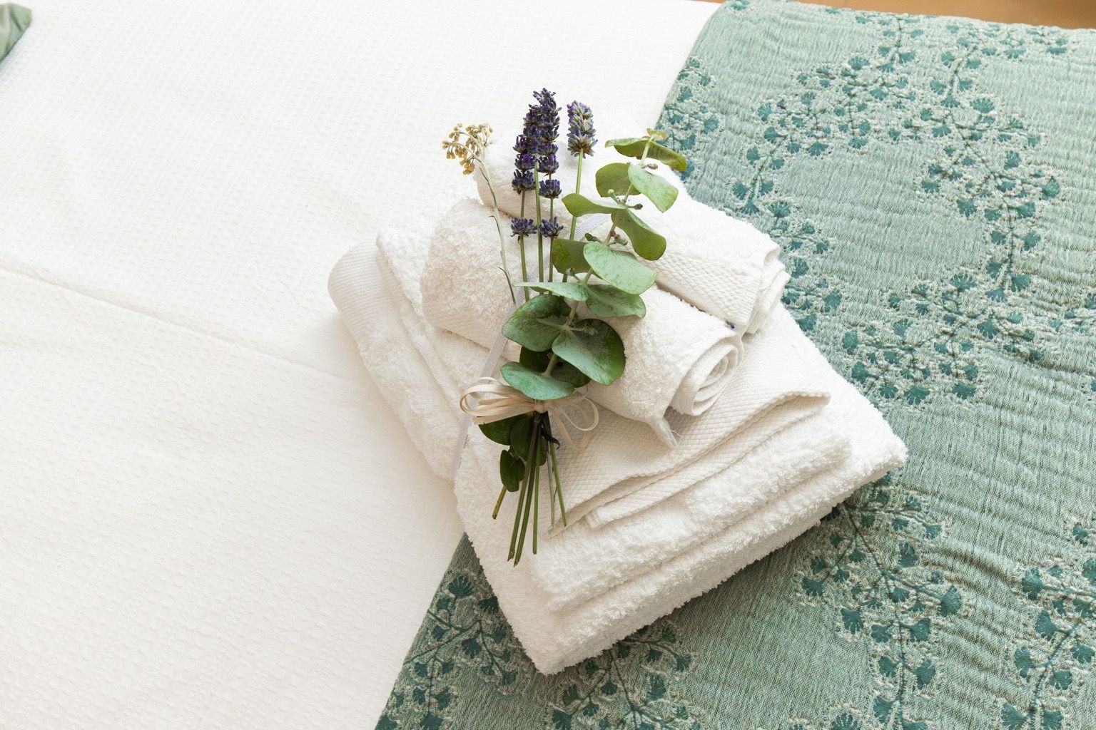 Ein ordentlich gefaltetes weißes Handtuch mit einem Zweig Lavendel und Eukalyptusblättern, platziert auf einem grün gemusterten Kissen.