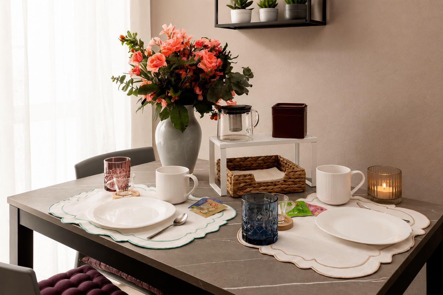 Une table à manger dressée avec des assiettes, des tasses, une cafetière, une bougie et un vase rempli de fleurs.