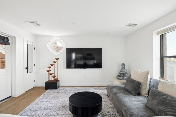 A modern living room with a flat-screen TV mounted on the wall, a black coffee table, a grey sofa with cushions, and a decorative wall piece.