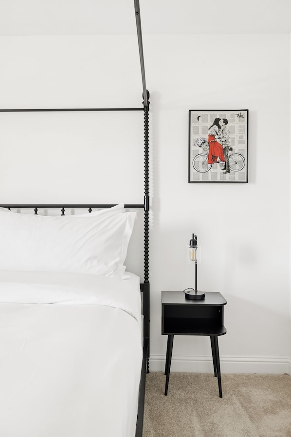 Un lit avec un cadre en métal noir, une literie blanche et une petite table de chevet noire avec une lampe.