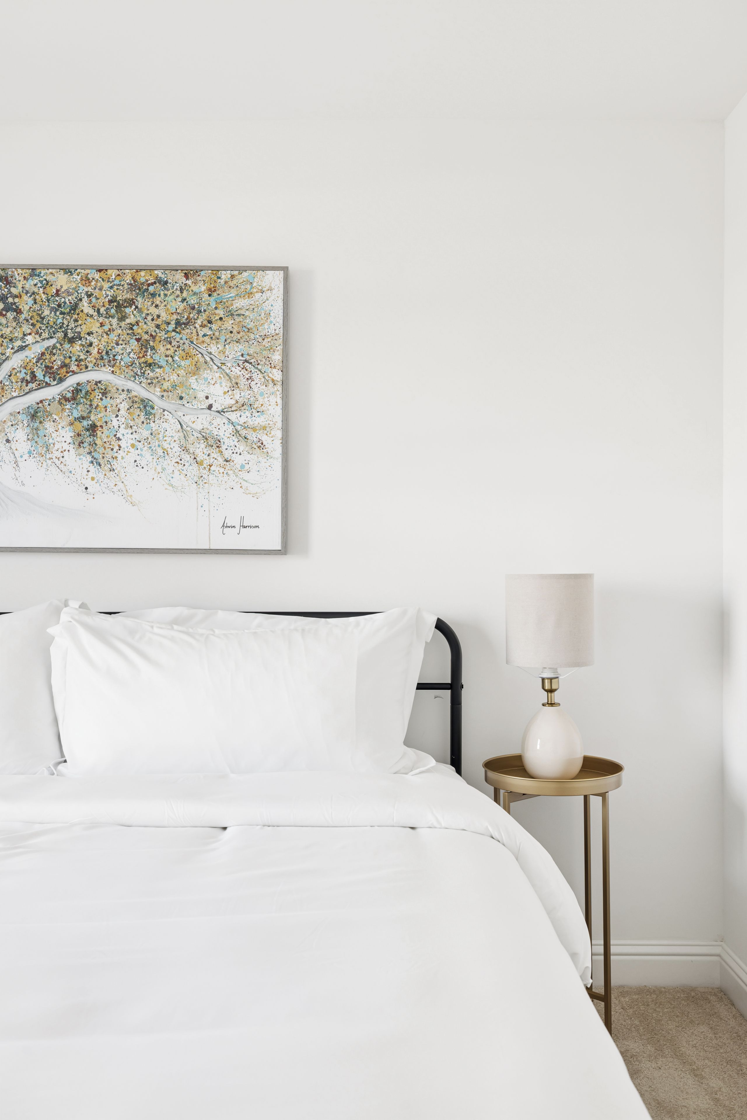 A neatly made bed with white linens, a small bedside table with a lamp, and a wall-mounted abstract painting.