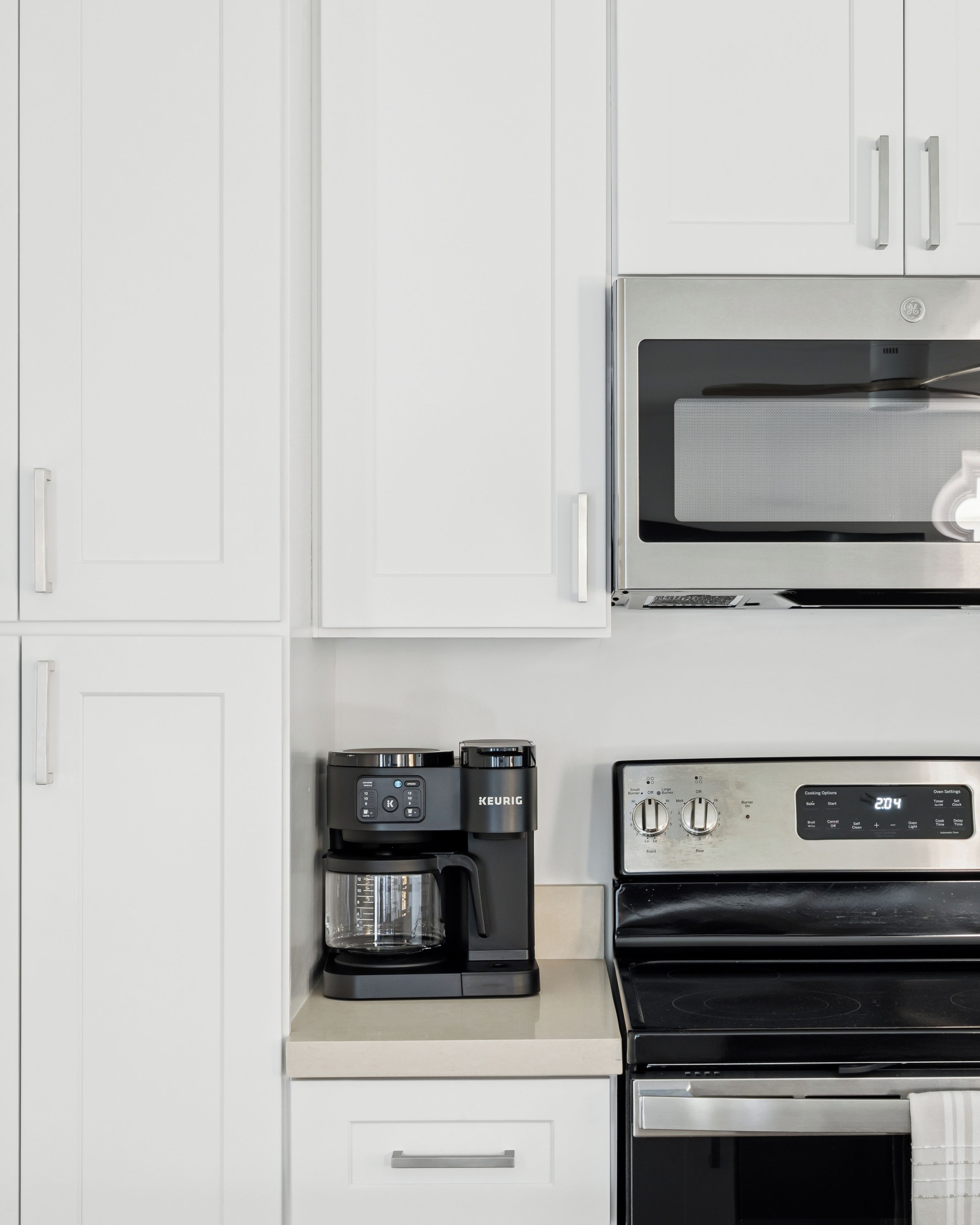 Una cocina con armarios blancos, un microondas GE, una cafetera Keurig y un horno de acero inoxidable.