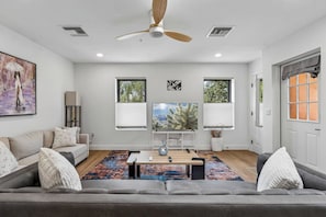 A modern living room with a grey sofa, a flat-screen TV, a ceiling fan, and a large window with blinds.