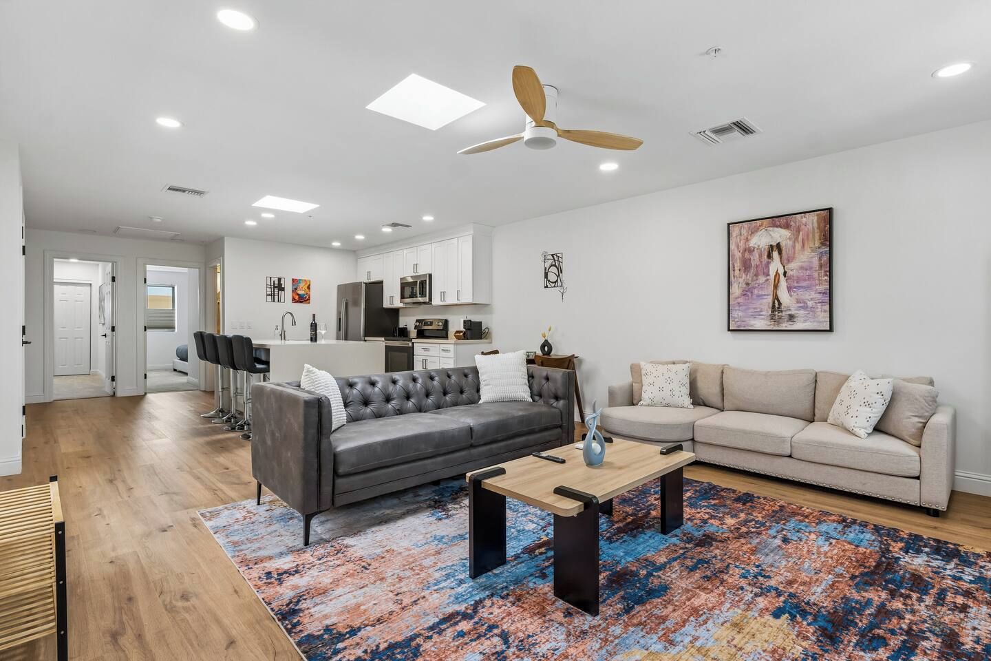 A modern living room with a grey sofa, beige sofa, and a coffee table. There is a kitchen in the background with white cabinets and a microwave.