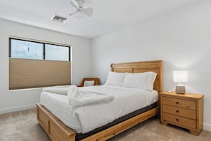 A bedroom with a wooden bed, a nightstand with a lamp, a window with blinds, and a ceiling fan.