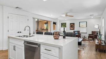 Una cocina moderna con una isla central, un comedor con sofá y sillas, y una sala de estar con televisor y lámpara.