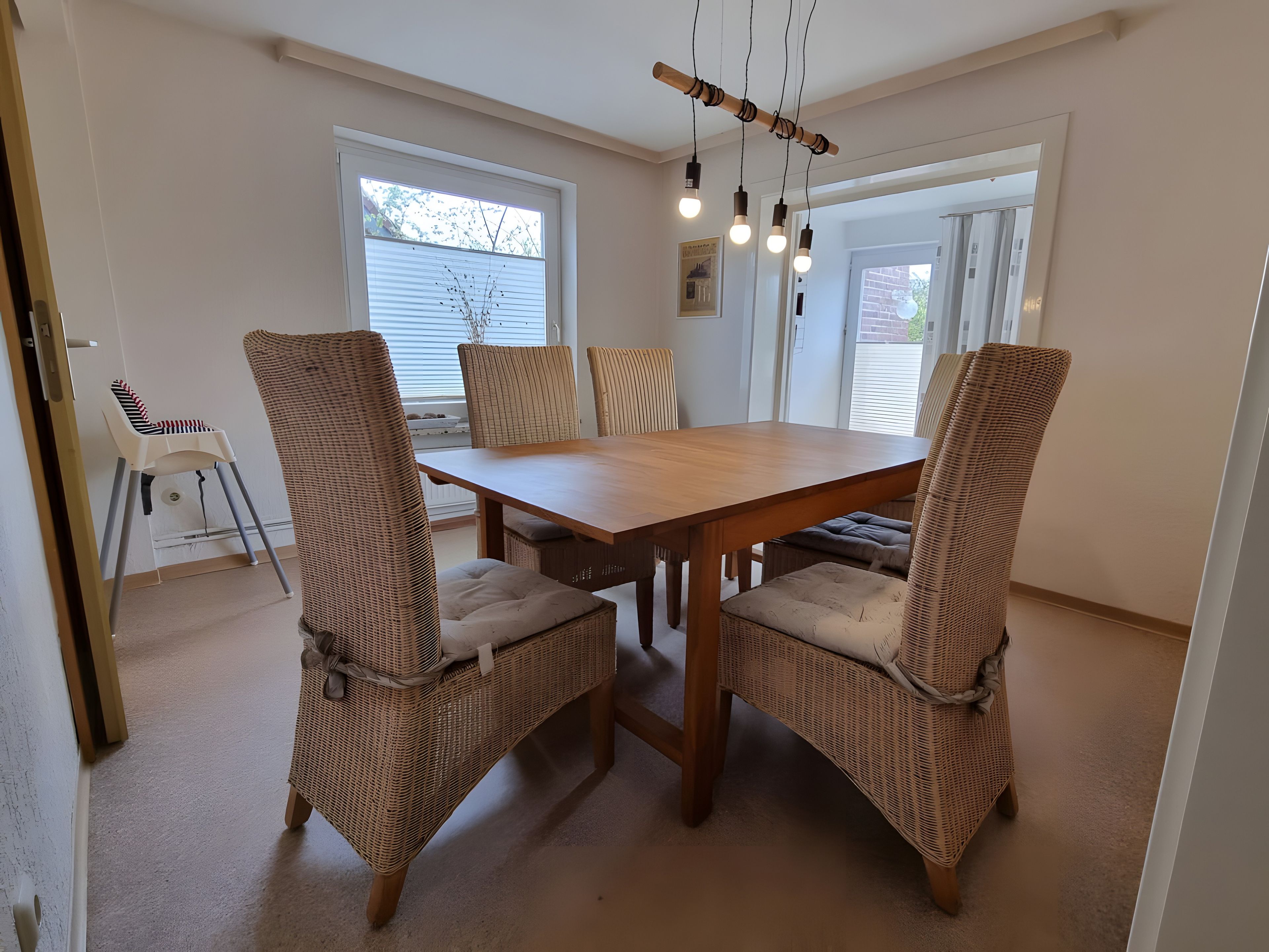 A dining area with a wooden table and wicker chairs, a high ceiling, and a view through a window.
