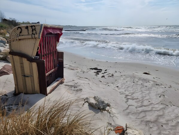 A beach chair with a woven backrest and a red seat, positioned on a sandy beach with waves in the background.