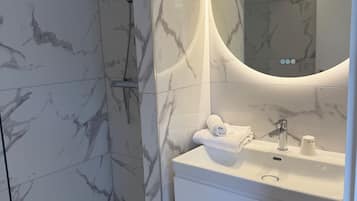 A modern bathroom with a glass-enclosed shower, a marble-tiled wall, a white vanity with a sink, and a round mirror.