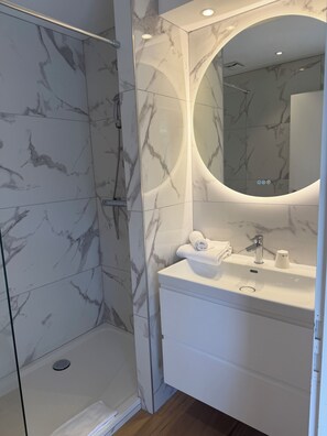 Een moderne badkamer met een douchecabine van glas, een wand betegeld met marmer, een witte wastafel met een wastafel en een ronde spiegel.