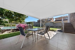 A covered patio area with a table and chairs, a pool in the background, and a brick wall. - Cosy Sunlit home with a Pool (Smithfield)