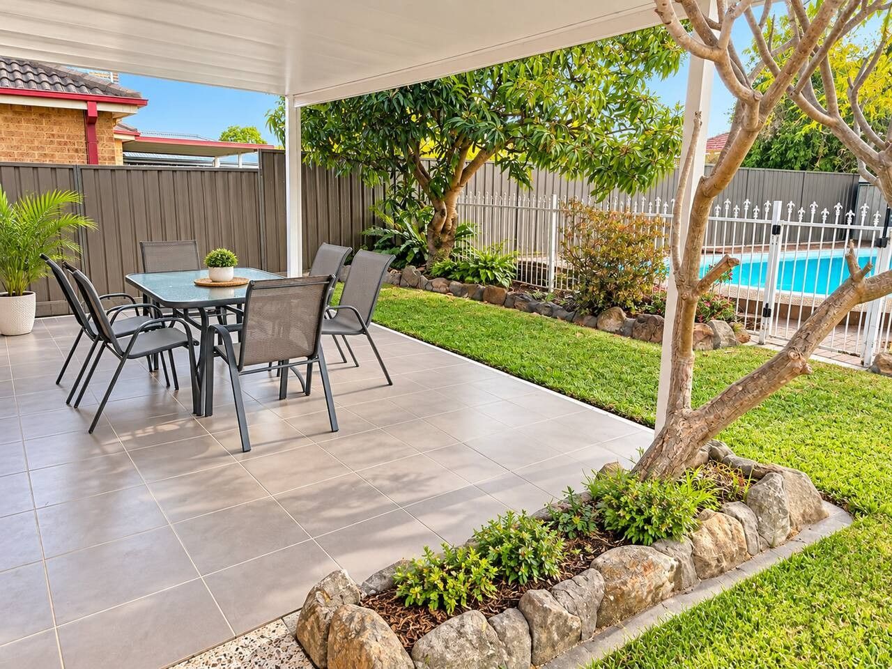 A covered patio with a table and chairs, a swimming pool in the background, and a fenced yard.