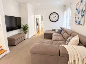 A living room with a flat-screen TV, a sectional sofa, a clock, and a painting. - Cosy Sunlit home with a Pool (Smithfield)