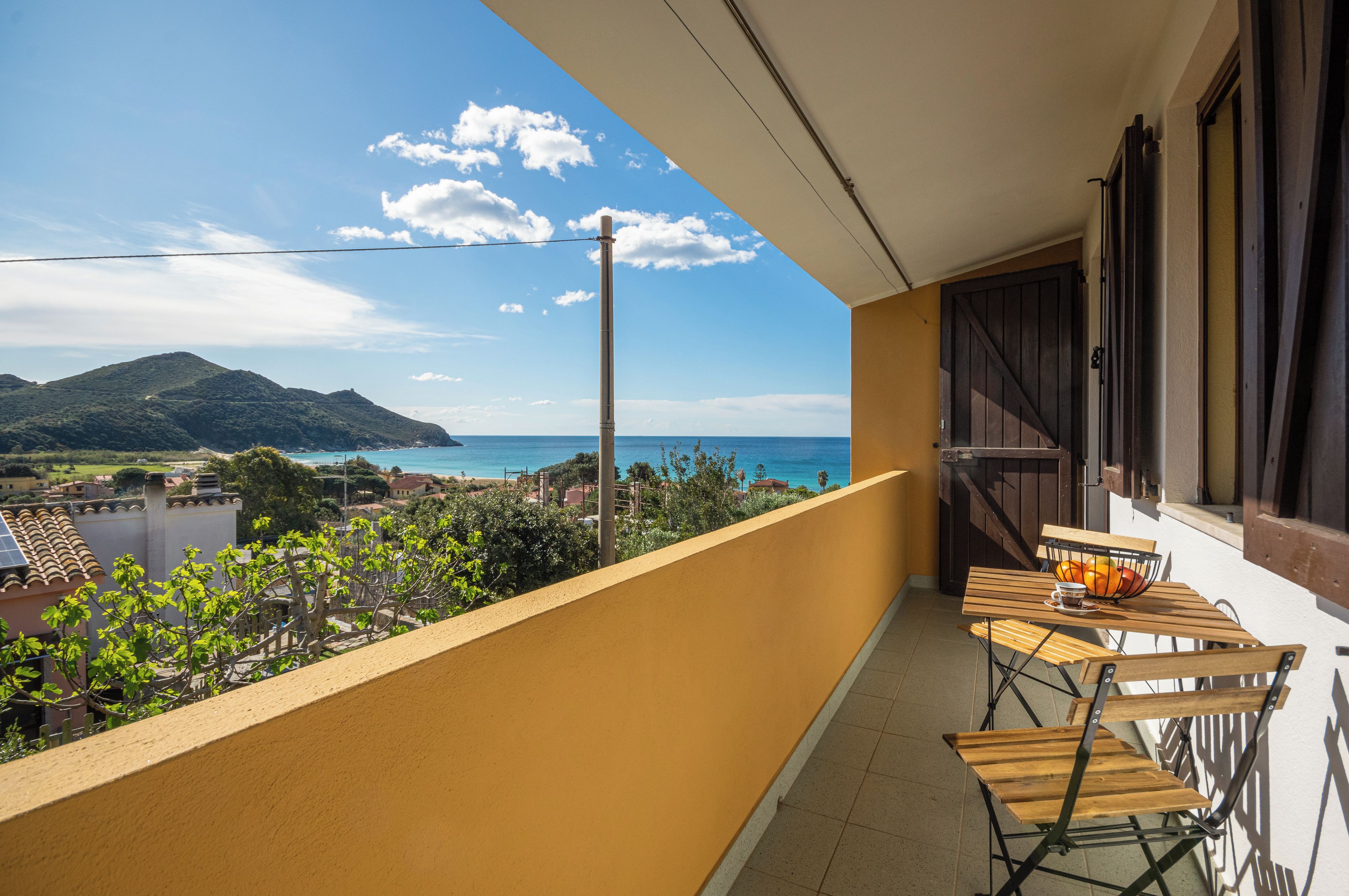 A balcony with a view of the sea and a mountain, a table with chairs, and a wooden fence.