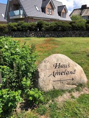 Ein Steinschild mit der Aufschrift 'Haus Ameland', umgeben von Grünpflanzen und einer Steinmauer.