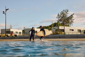 Two people in wetsuits walking in shallow water with surfboards, a streetlight, and buildings in the background.