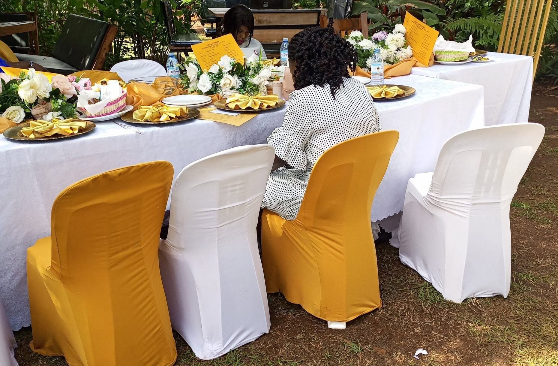 Mesa colocada al aire libre con sillas blancas y amarillas, un menú y arreglos florales.