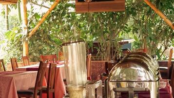 An outdoor dining area with tables set for a meal, surrounded by greenery and a wooden structure overhead.