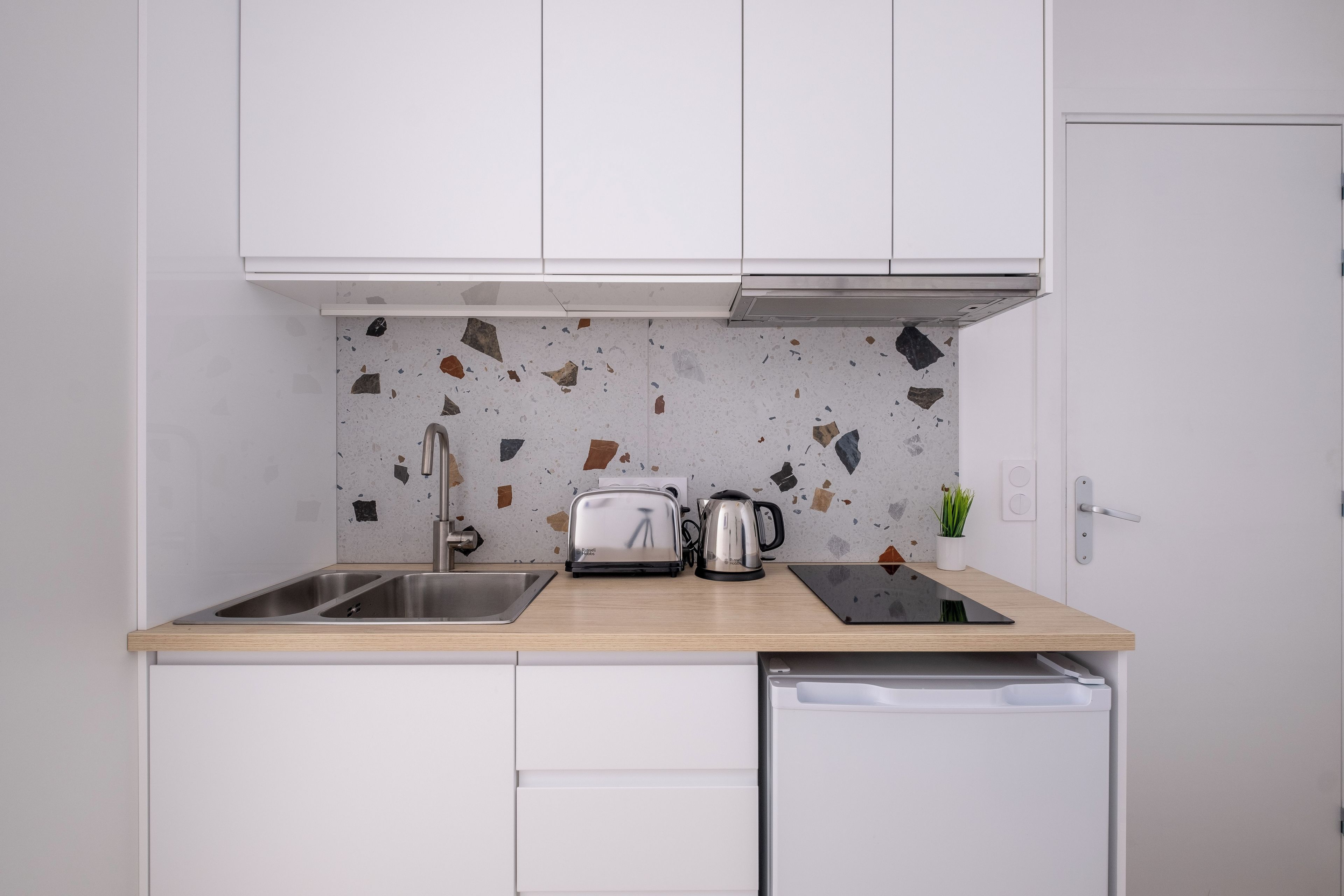 Una cocina con armarios blancos, una encimera, un fregadero, un tostador, una tetera y una pequeña planta en maceta.