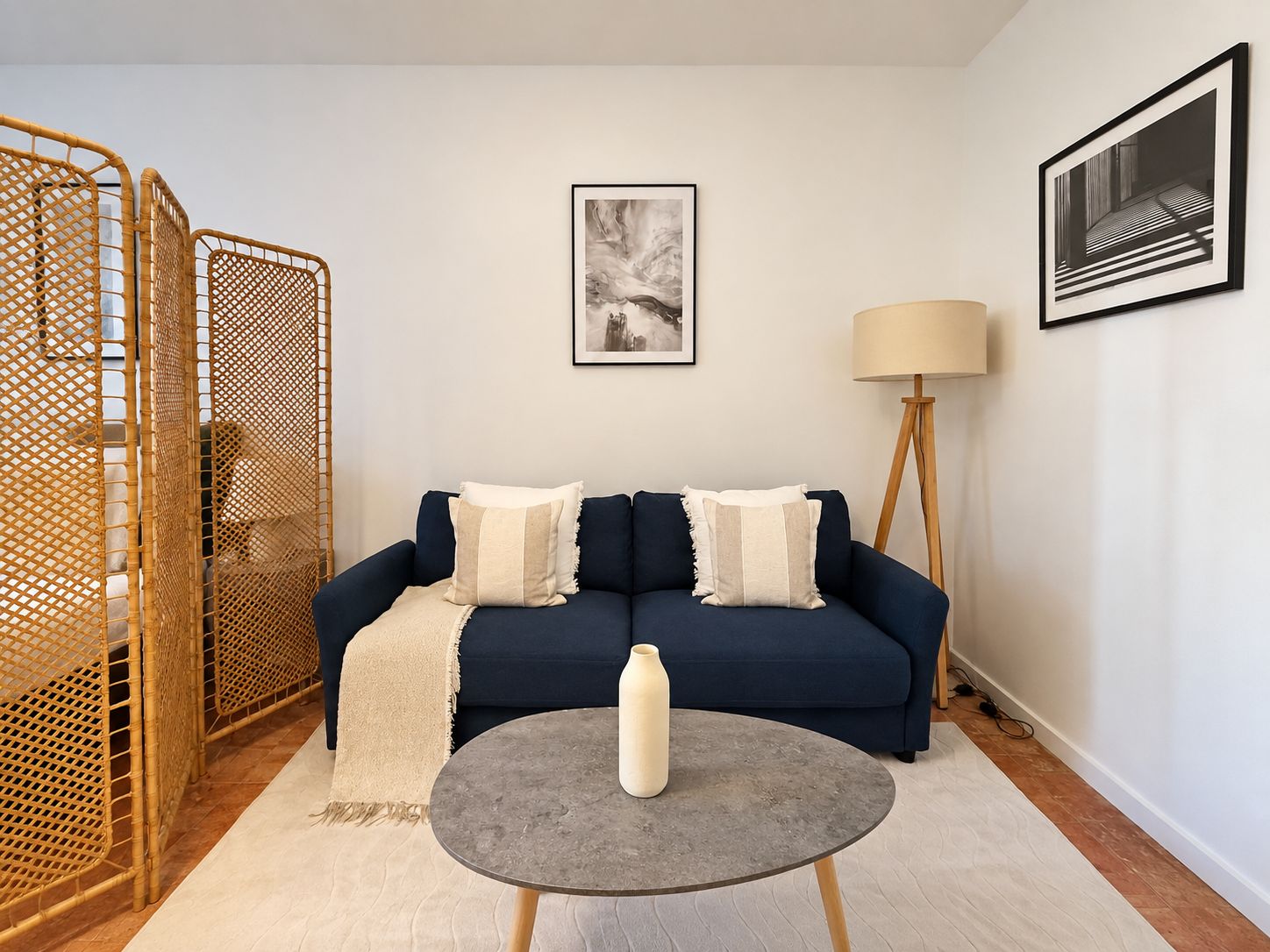 Un salón con un sofá azul, dos almohadas, una mesita redonda con una botella, una lámpara de pie y un cuadro en la pared.