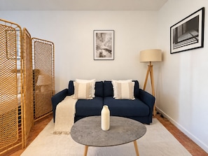 Un salón con un sofá azul, dos almohadas, una mesita redonda con una botella, una lámpara de pie y un cuadro en la pared.