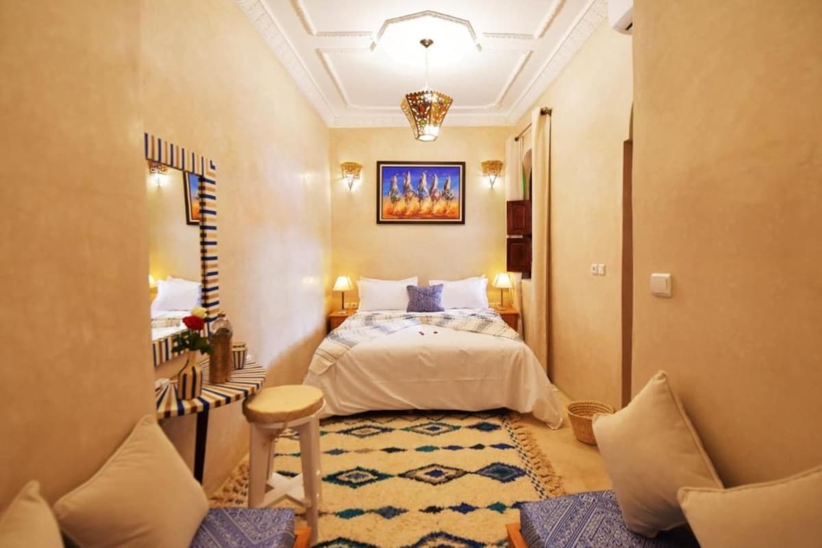 Una habitación de hotel con cama, mesitas de noche, una silla, una alfombra y un cuadro en la pared.