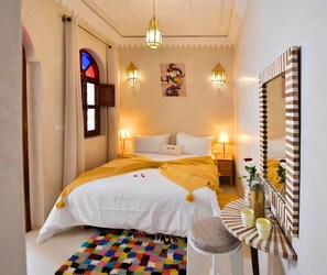 Una habitación de hotel con cama, lámparas de noche, alfombra colorida, una mesita con un florero y un espejo.