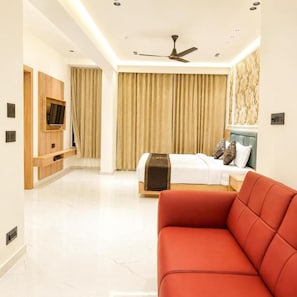 A hotel room with a bed, a red sofa, a ceiling fan, a TV mounted on the wall, and a window with curtains. - Hotel Mangla Inn (Dwarka)