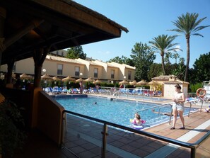 Ein Swimmingpool mit Liegestühlen und Sonnenschirmen, umgeben von Palmen und einem Gebäude mit einem Ziegeldach.