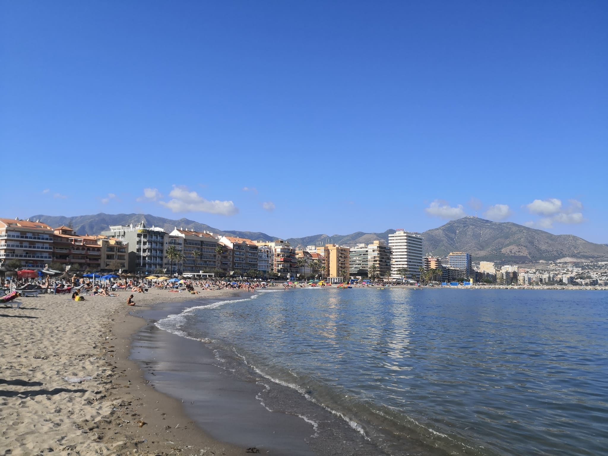 Une plage avec des bâtiments en arrière-plan, un ciel dégagé et des montagnes à l’horizon.