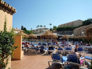 A poolside area with lounge chairs and umbrellas, surrounded by buildings and palm trees. - Luxe Duplex Appartement in Marina del Sol, Mijas (Mijas)