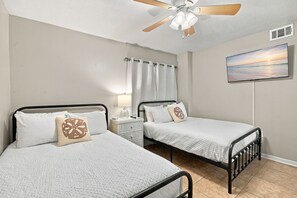 Une chambre avec deux lits, un ventilateur de plafond, une table de chevet, une lampe, un téléviseur et un tableau représentant un coucher de soleil.