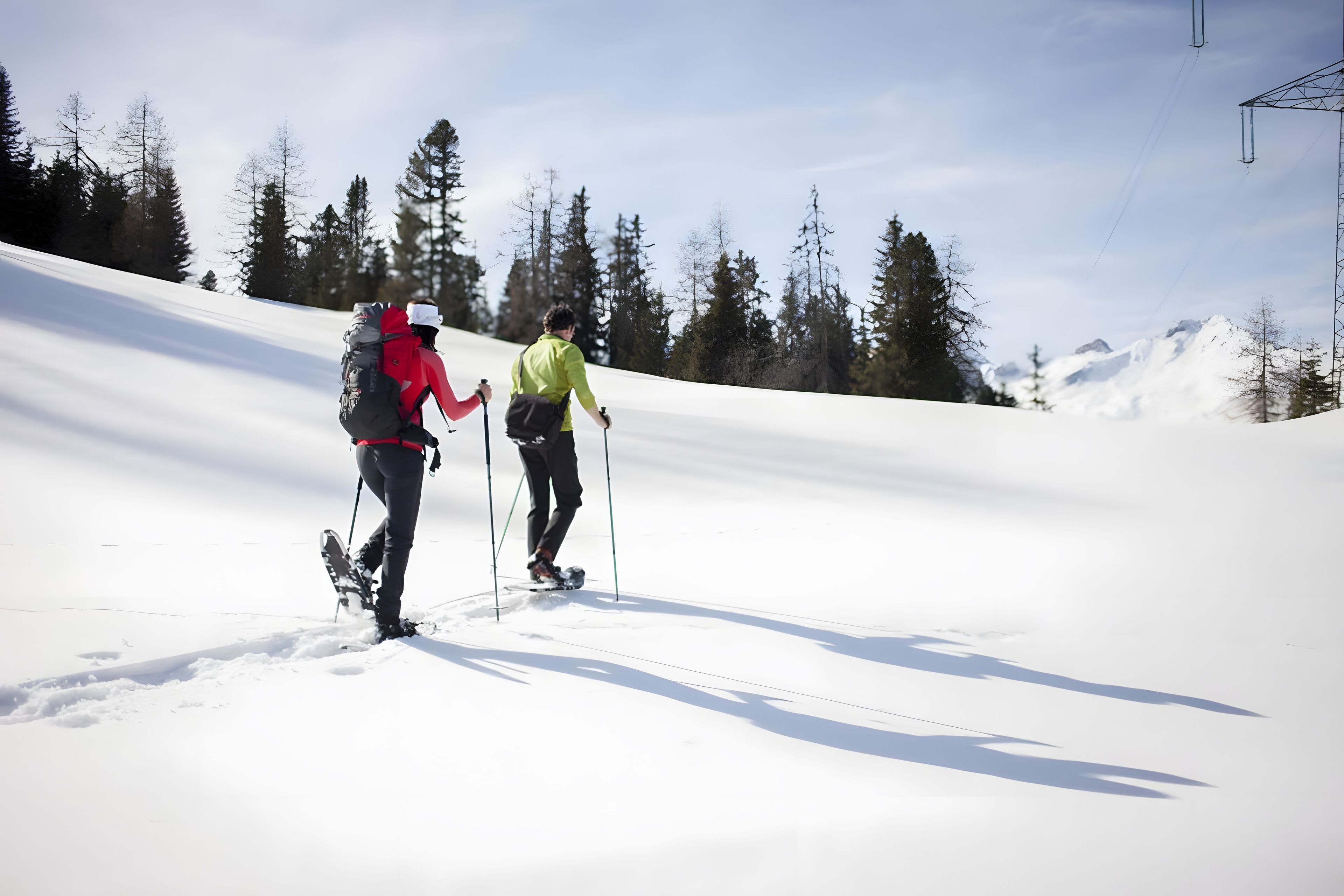 Duas pessoas fazem caminhada em uma encosta coberta de neve, com árvores ao fundo.