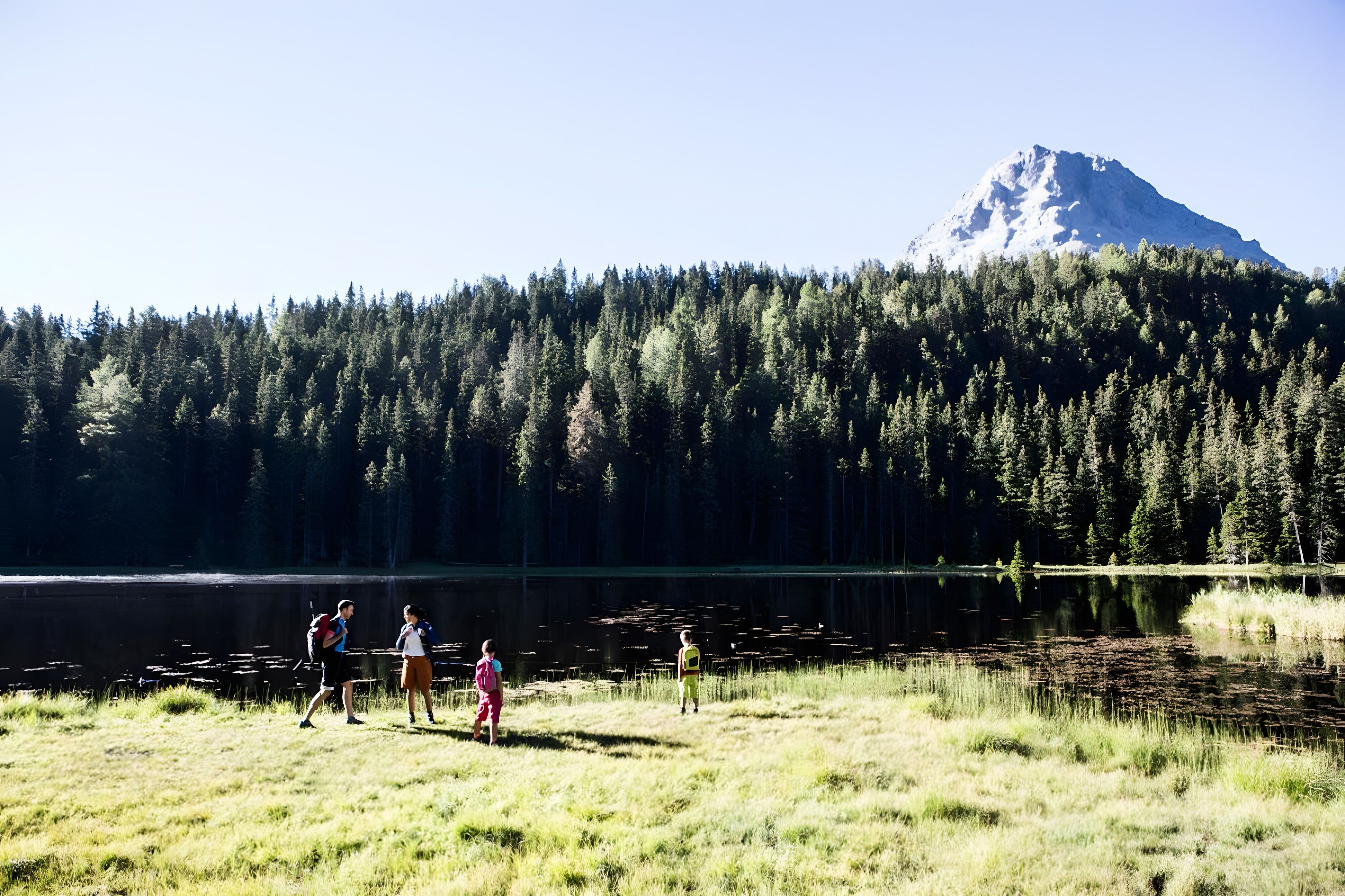 A group of people walking near a lake with a forest and a mountain in the background.