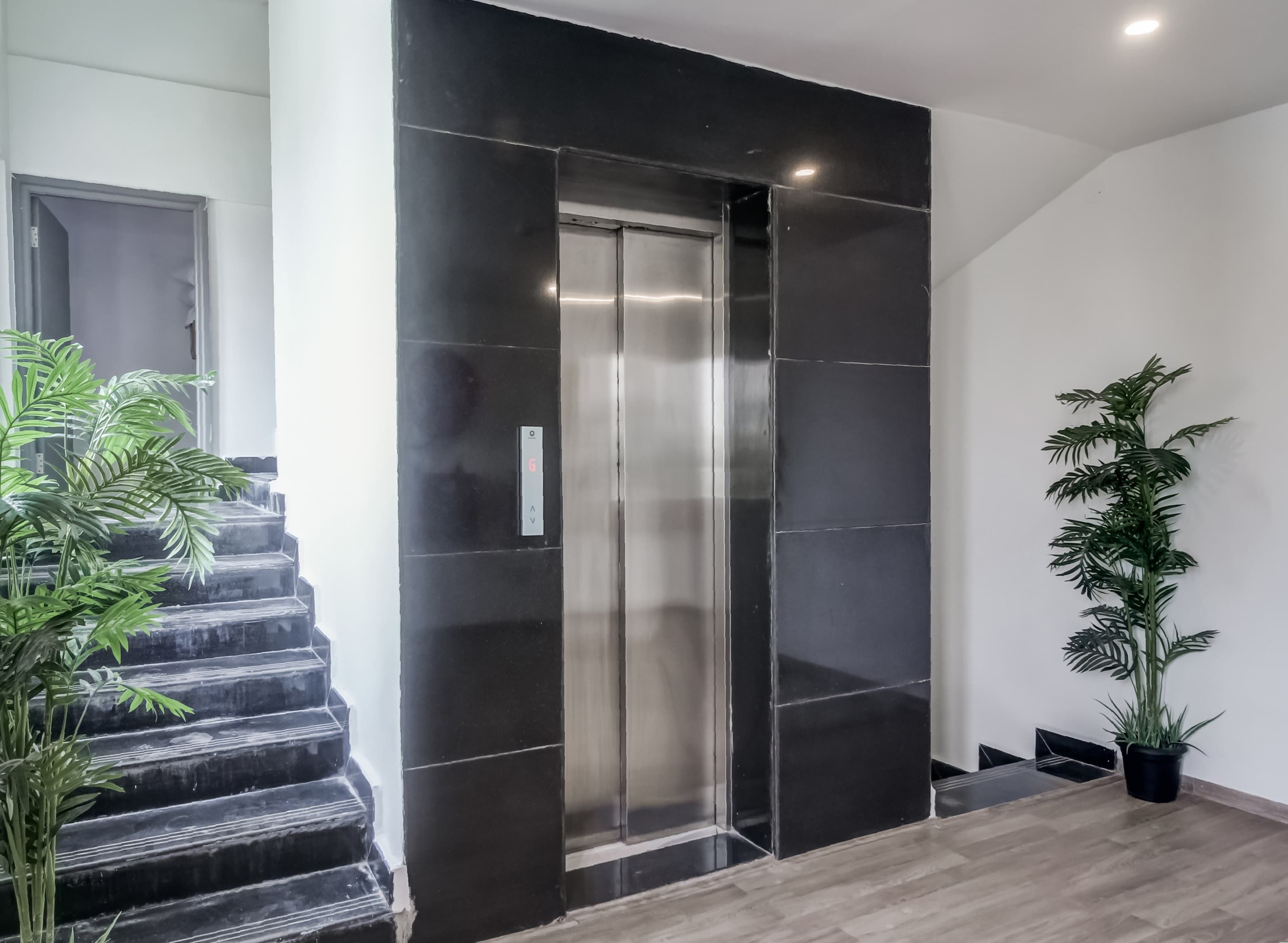 Un ascensor moderno con detalles metálicos, flanqueado por paredes con azulejos negros y suelo de madera.