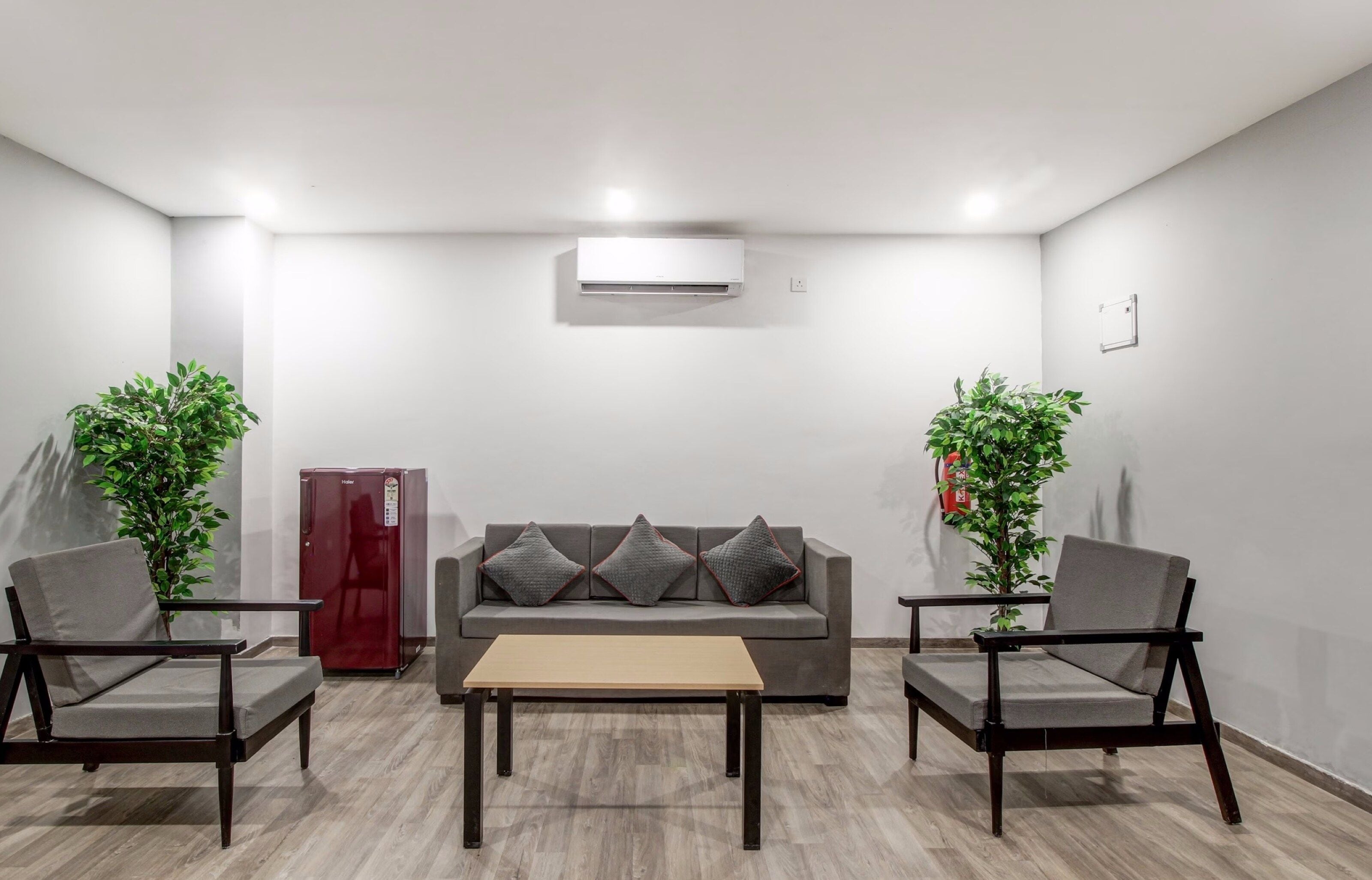 Een moderne woonkamer met een grijze bank, twee fauteuils, een houten salontafel, een rode koelkast en twee potplanten.