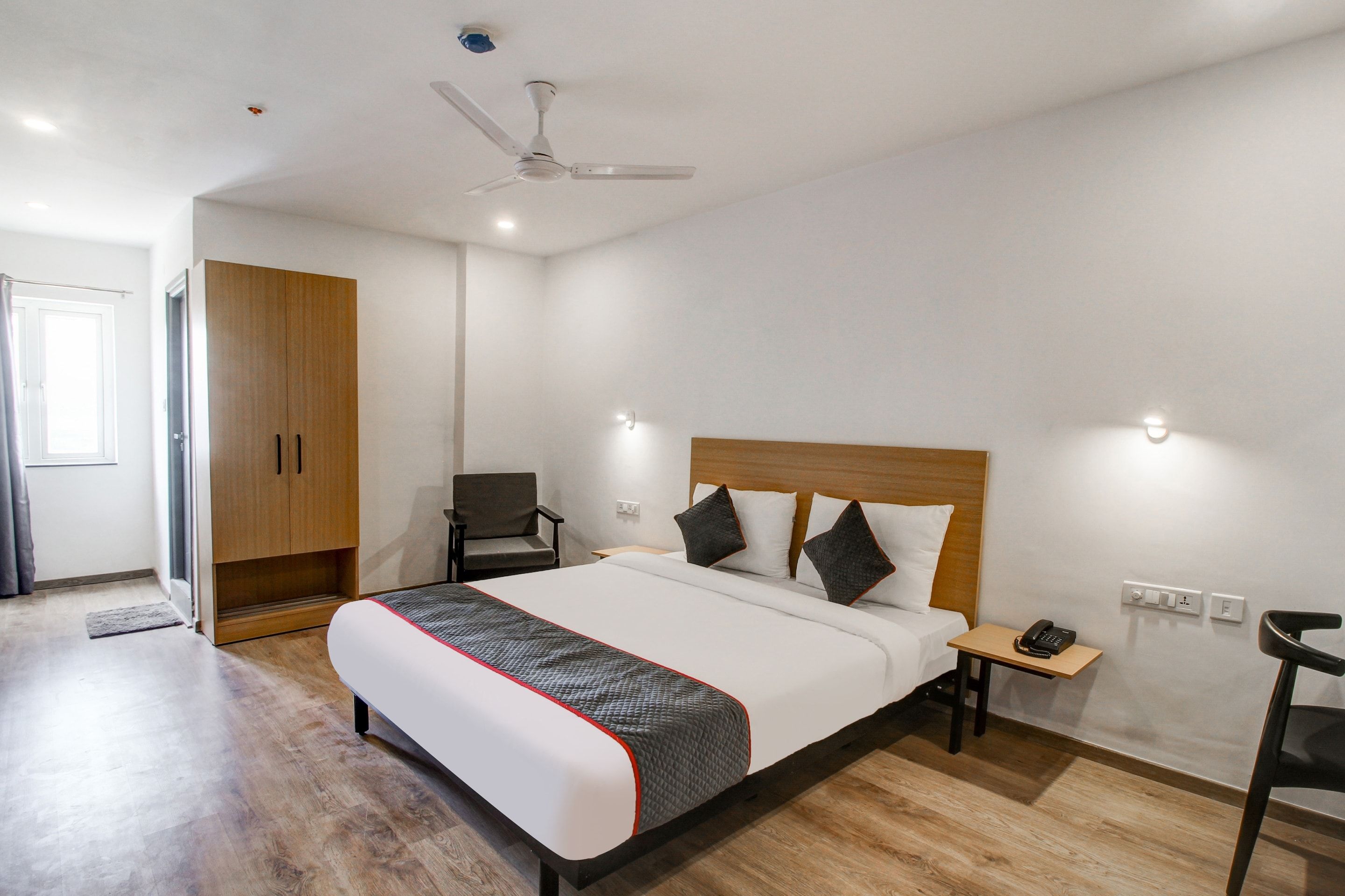 A hotel room with a bed, a chair, a desk, a wardrobe, and a ceiling fan.