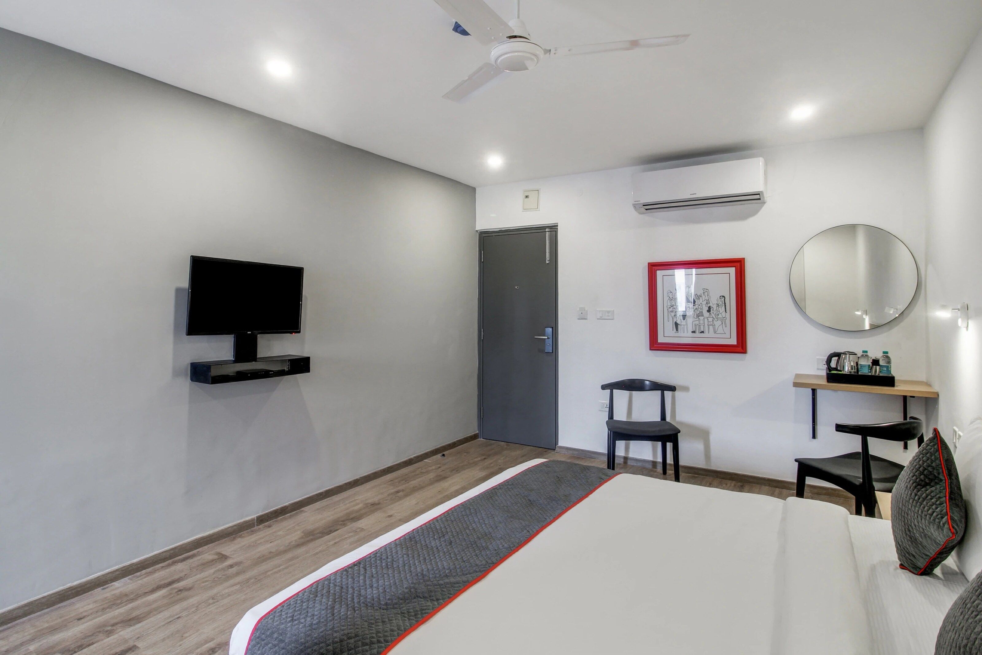 A hotel room with a bed, a flat-screen TV mounted on the wall, a desk with a chair, a mirror, and an air conditioning unit.