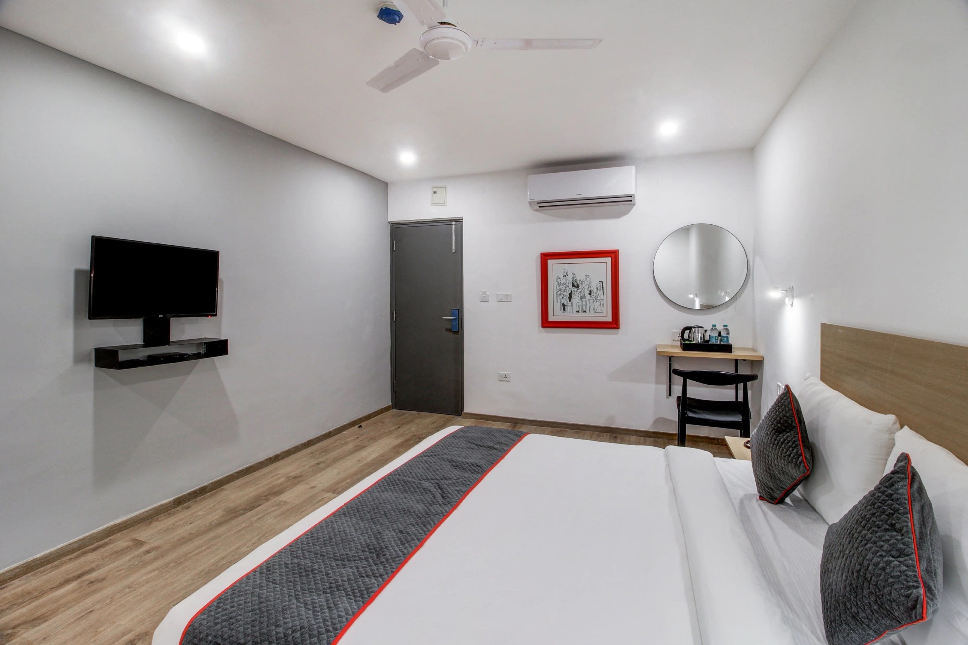A modern hotel room with a flat-screen TV, a bed with pillows, a bedside table with a mirror, a desk with a chair, and a door.